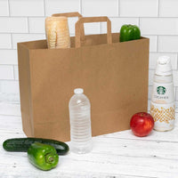 Brown paper grocery bag with flat handles filled with groceries, including a bottle of water, cucumber, and bread, on a white wooden table.