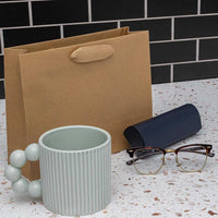 Medium brown paper bag with ribbon handles, a ceramic mug, eyeglasses, and a glasses case on a kitchen counter.