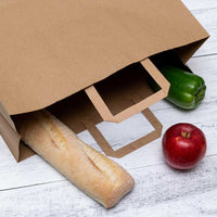 Large flat handle paper bag with a baguette, green bell pepper, and red apple on a white wooden surface.